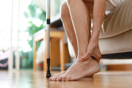 Adult woman sitting on sofa holds her ankle due to pain