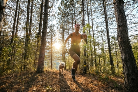 Young woman jogging on a wooded trail in spring with her dog tagging along behind her.