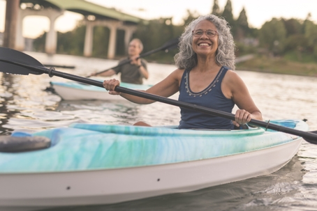 Older woman smiling at the camera as she kayaks with her husband on a beautiful day at the river. | ProFysio Physical Therapy